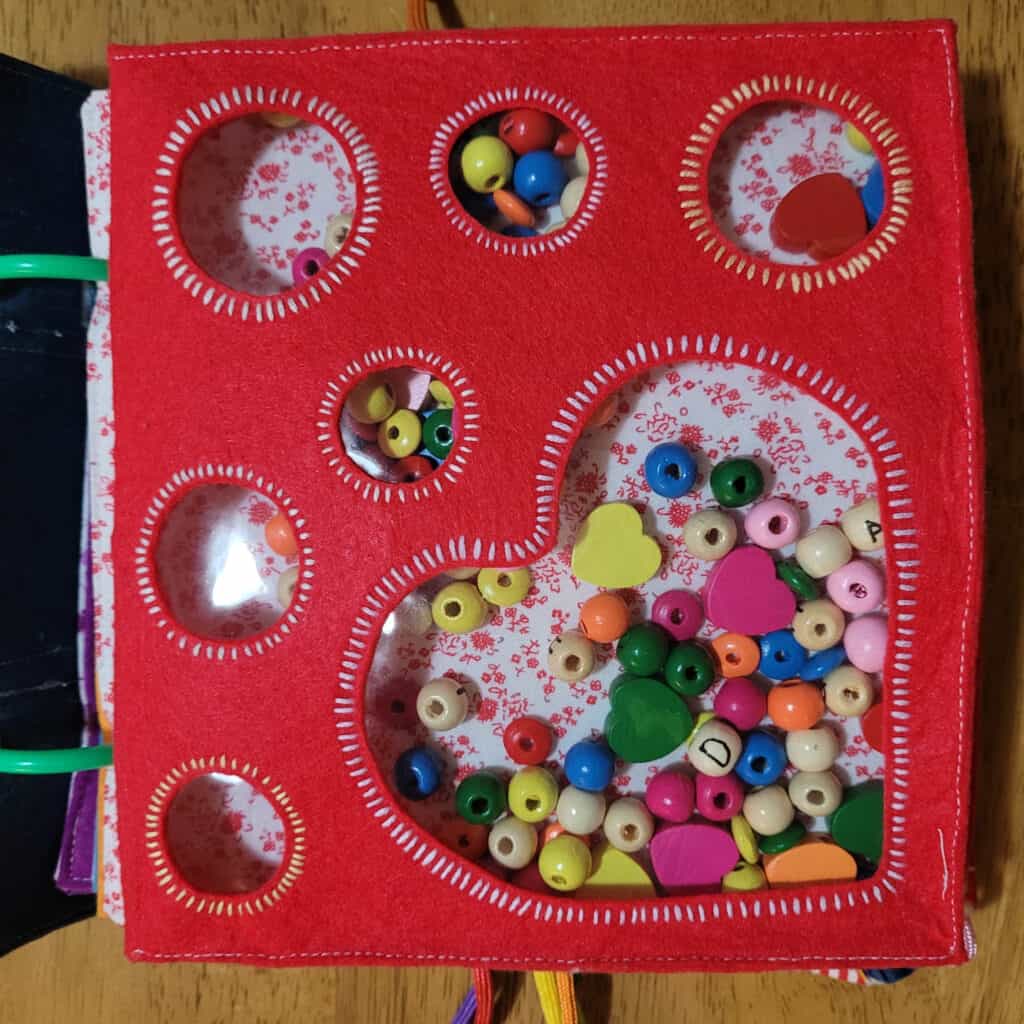 A busy book page. It is red felt, with a heart and circles cut out and covered with clear plastic. Behind the clear plastic is a background of white fabric with red flowers. It is filled with beads in red, yellow, orange, pink, blue, green, magenta and natural wood colours. The beads are mostly round, with some flat shapes and some heart shapes. The plain wood ones have letters drawn on them.