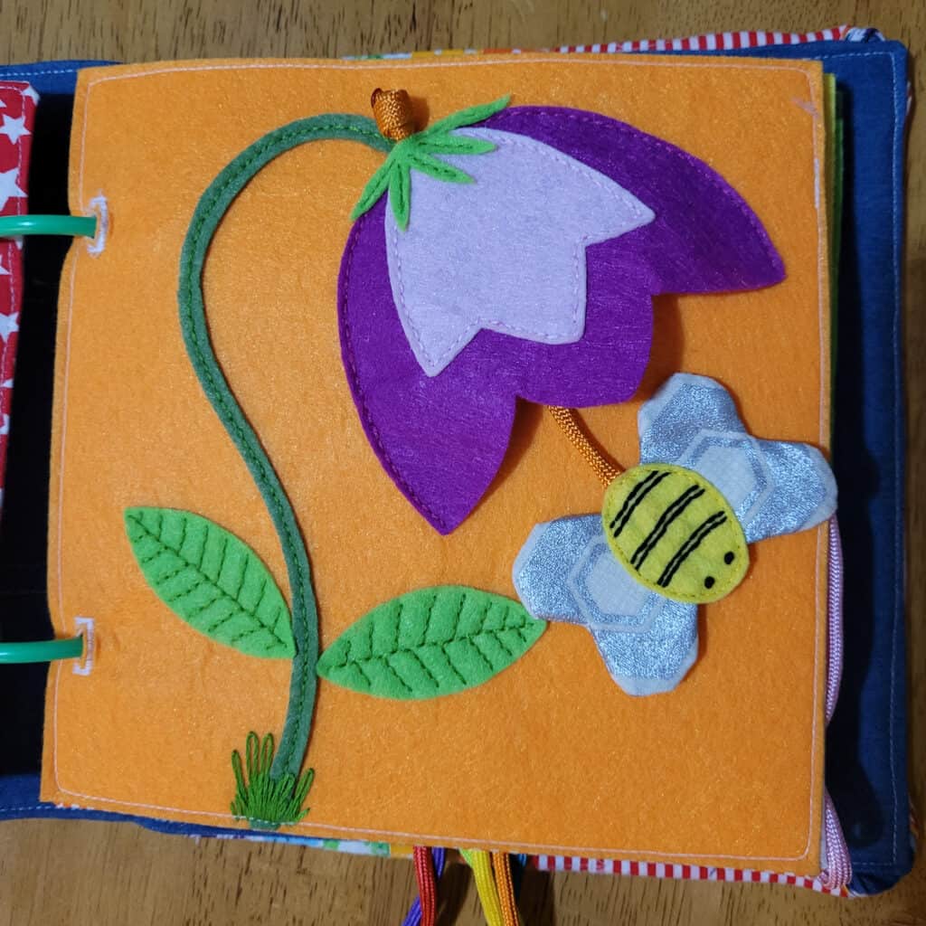 A busy book page with an orange background. It has a large purple and pink flower with a green stem and leaves. There is a sewn bumble bee that can be pulled in and out of the flower on a cord.