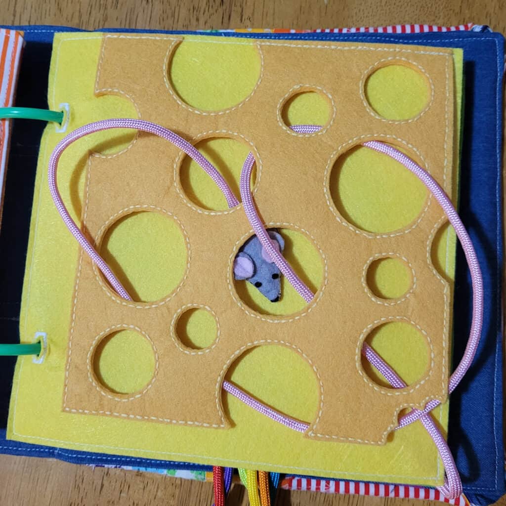 This busy book page has a yellow background with a sort of yellowy-orange flap on top, which is shaped like a piece of Swiss cheese, with holes cut out. There is a felt mouse with a very long pink tail, which can be woven through the holes.