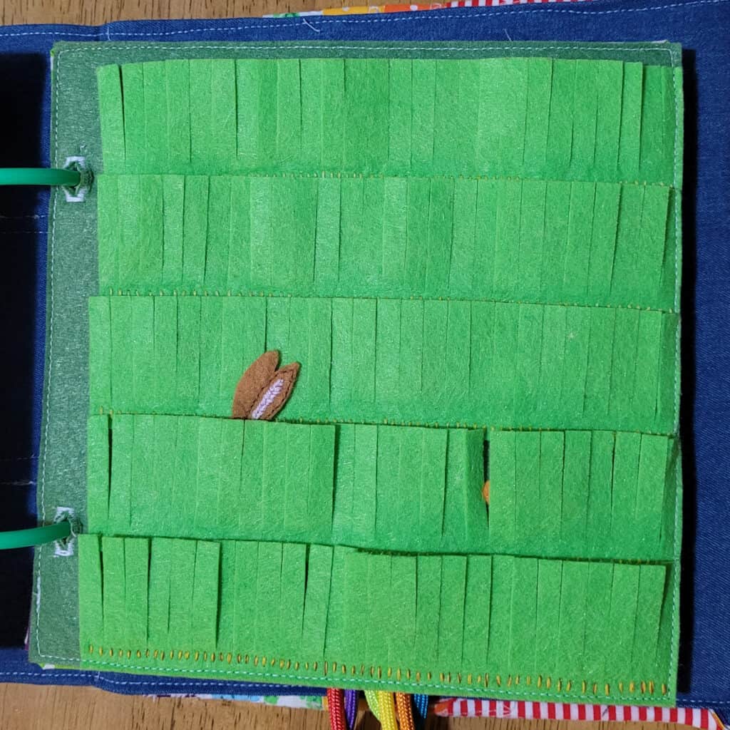 A busy book page. It has a dark green background and bright green strips of felt cut to look like grass. There are bunny ears sticking out from behind one of the rows of grass.