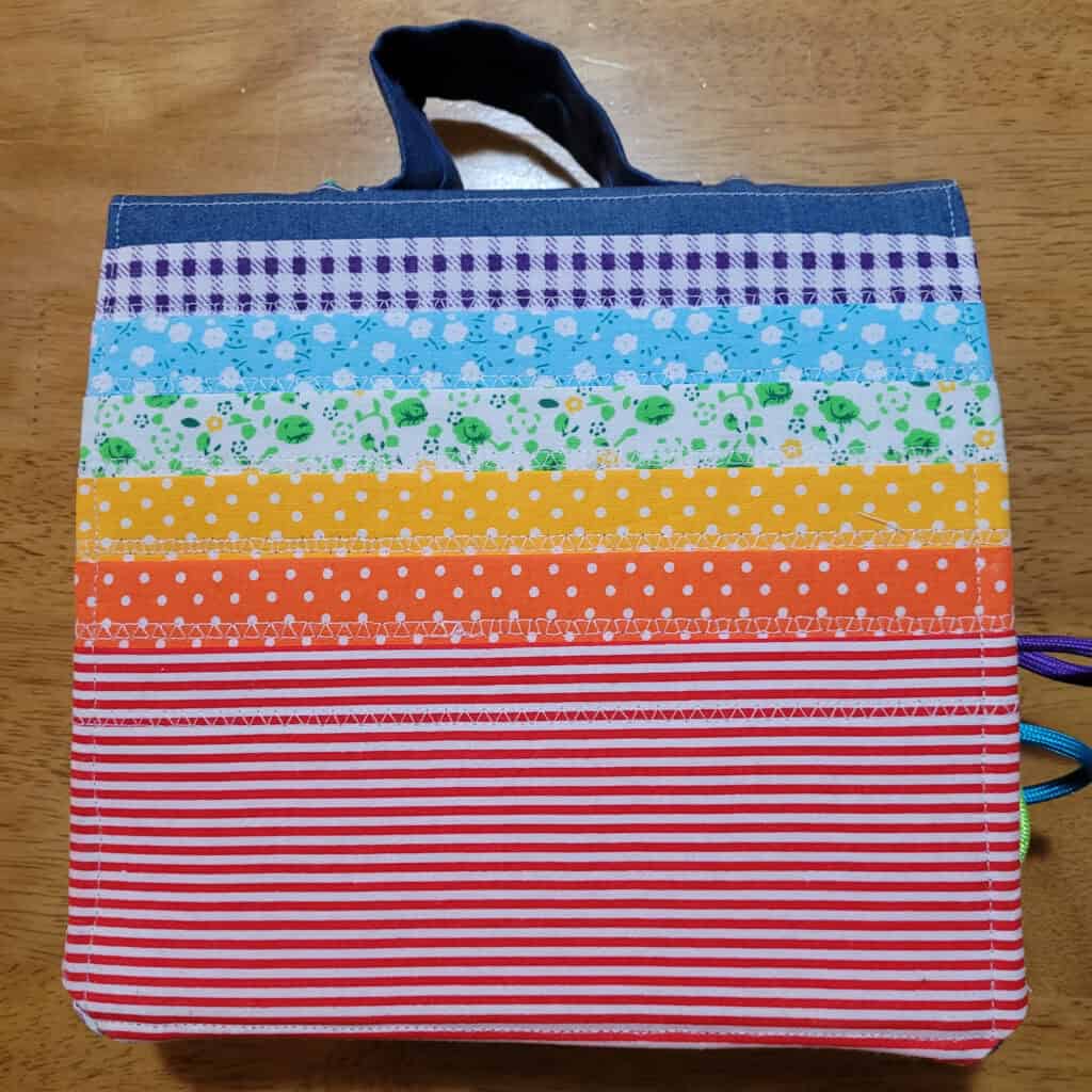 The cover of a busy book. You can see denim at the top, with a denim handle, and pockets in various colours and patterns, forming a rainbow. There are some cords coming out the right side of the book.