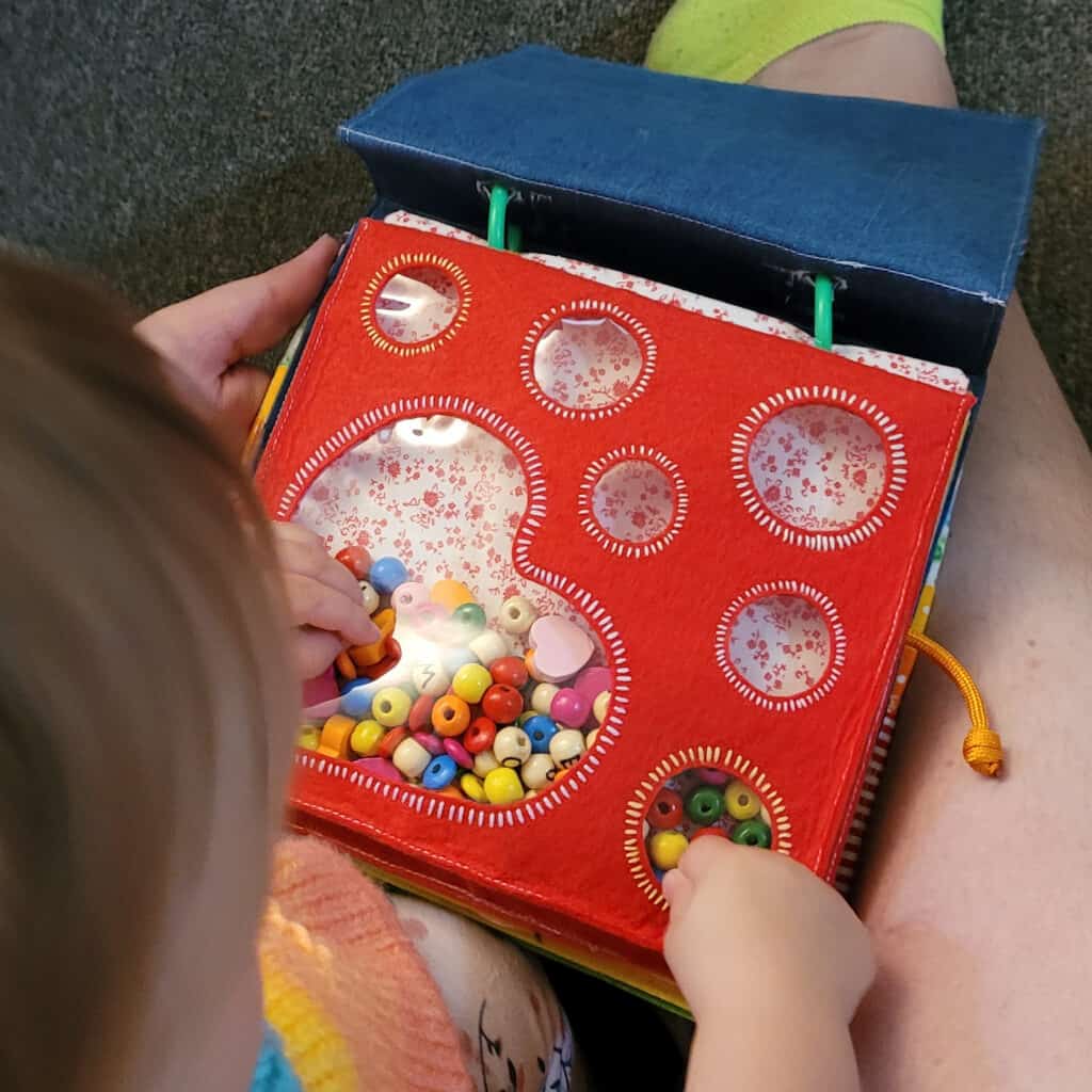 A busy book page. It is red felt, with a heart and circles cut out and covered with clear plastic. Behind the clear plastic is a background of white fabric with red flowers. It is filled with beads in red, yellow, orange, pink, blue, green, magenta and natural wood colours. The beads are mostly round, with some flat shapes and some heart shapes. The plain wood ones have letters drawn on them. We can see the top of a small child's head in the bottom left corner, and the child's hands pointing at the page. The child appears to be sitting on an adult's lap, we can see one leg on the right side of the image, partially under the book, with a neon green sock at the top, and an adult hand supports the busy book on the left side.