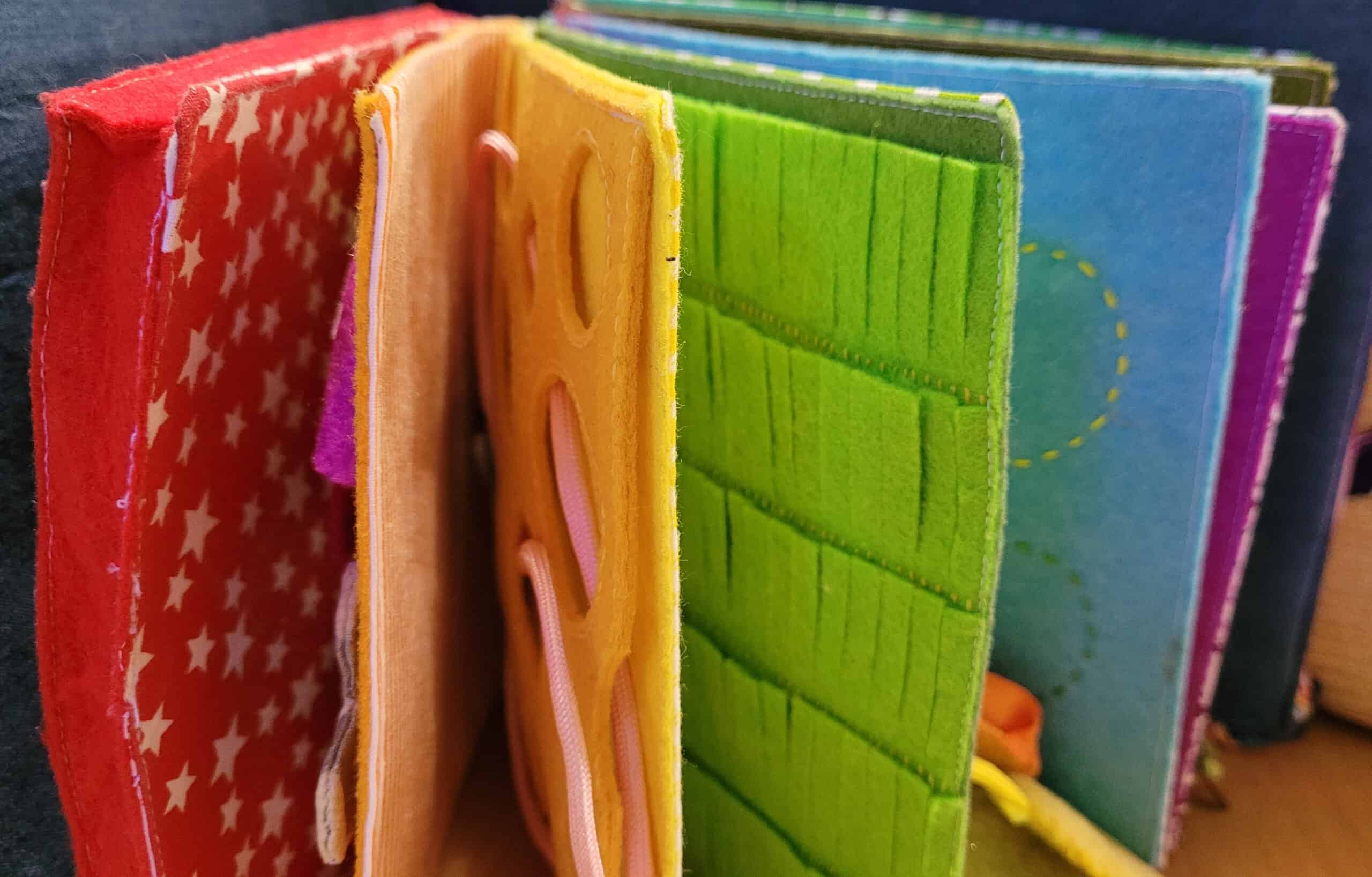 Felt busy book pages. They are bound together on one side, and are in rainbow colours left to right. Some details can be seen, like holes cut out on the yellow page and grass shapes on the green page.