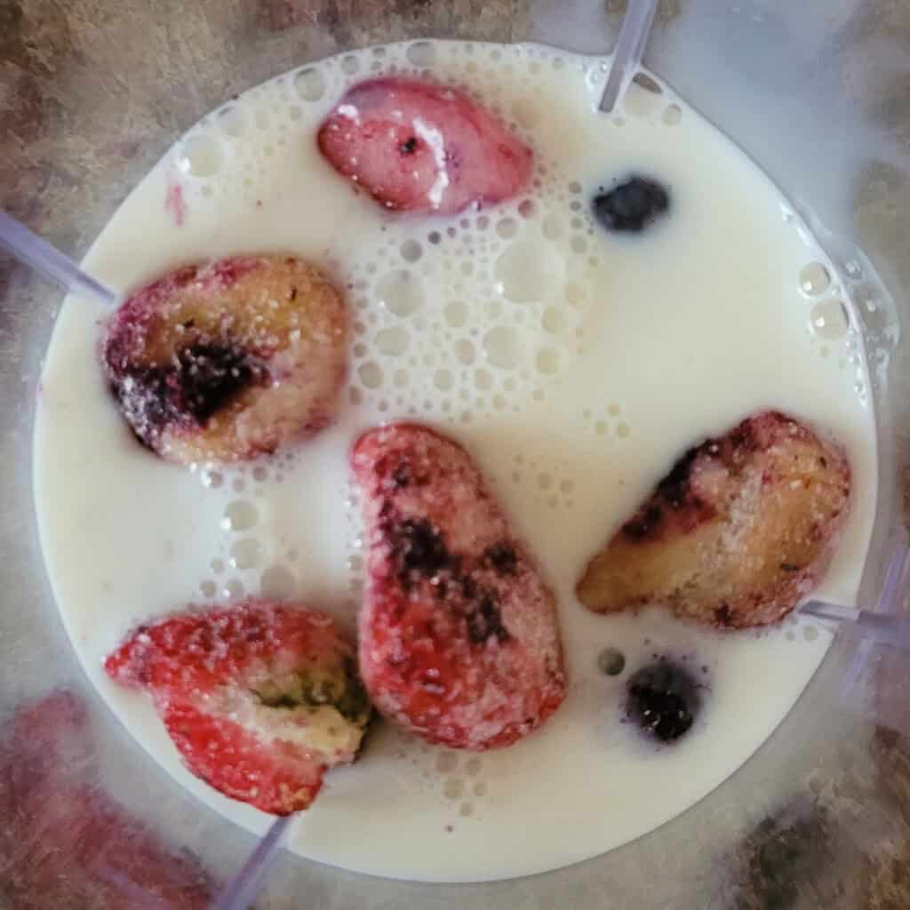 Frozen strawberries, bananas and blueberries with milk in the bottom of a blender.