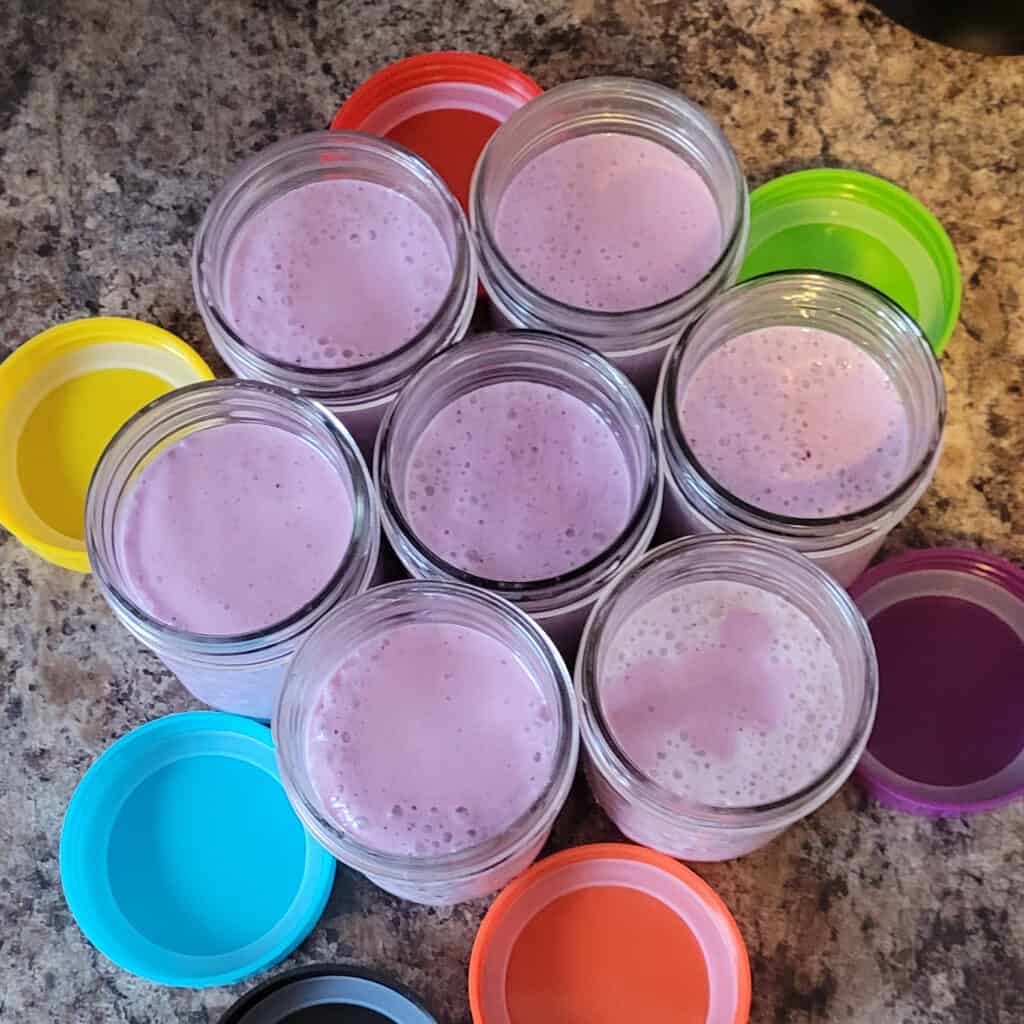 A top view of 7 half pint mason jars filled with light purple-ish yogurt drinks. There are colourful plastic lids on the counter around them.
